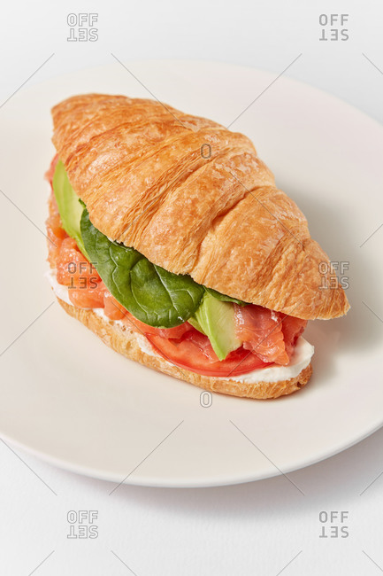 Close up view of freshly cooked homemade croissant sandwich with natural salmon fish, avocado, green leaf and ripe tomatoes on a ceramic plate on a white background, copy space.