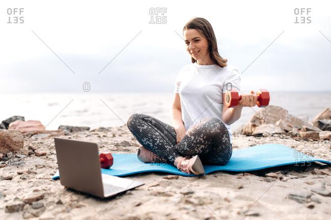 Fitness workout by video tutorial. A young girl does exercises with dumbbells while sitting on a mat and watches a video fitness lesson during a workout in nature