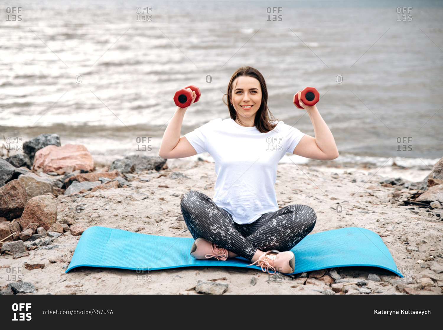 Outdoor workout. Young caucasian smiling girl gets fit. She sits on a mat in sportswear and doing exercises with dumbbells in a good mood
