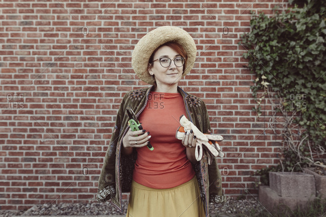 Portrait of mature woman with gardening gloves and pruner standing in front of brick wall