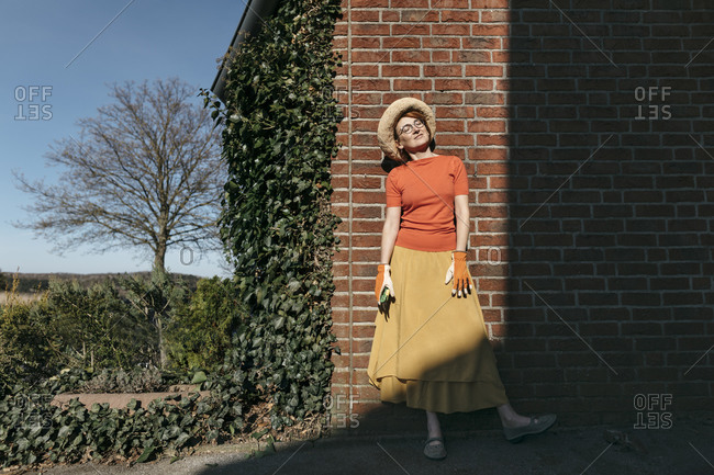 Portrait of mature woman with gardening gloves and pruner standing in front of brick wall enjoying sunlight
