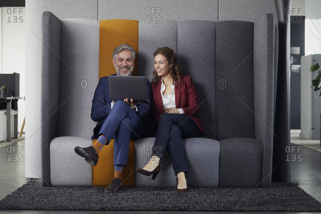 Happy businesswoman and businessman sitting on couch in office sharing laptop