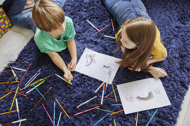 High angle view of boy and girl lying on blue rug, drawing with coloring pencils.