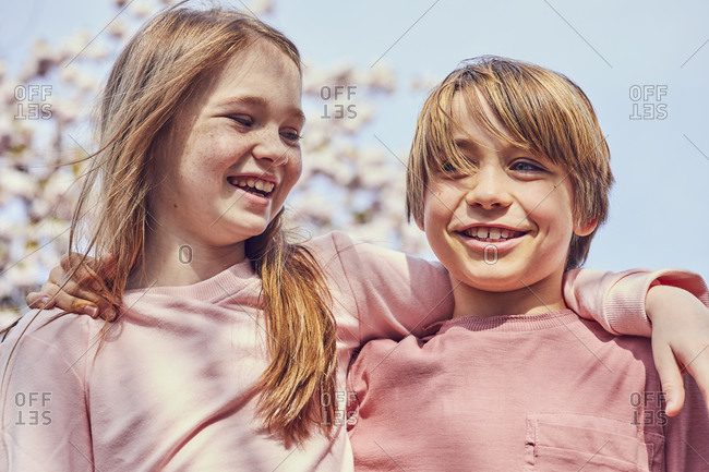 Smiling brunette boy and girl standing outdoors, arms around shoulders.