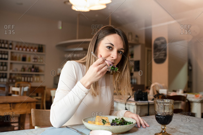 A woman seated at a cafe table eating a dish of vegan food, vegetables on her fork.