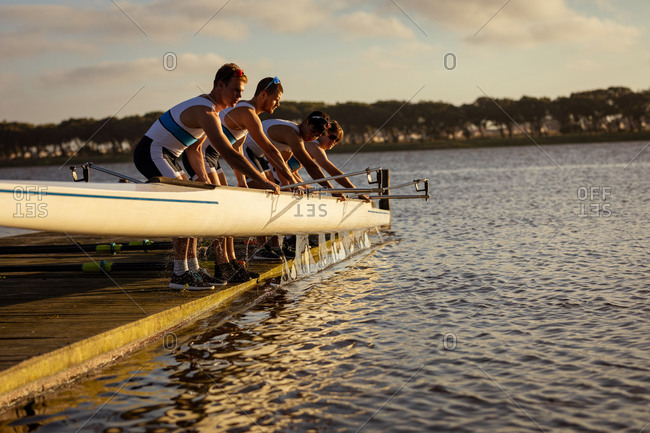 Side view of a rowing team of four Caucasian men taking a boat out of water standing on a jetty on the river at sunset