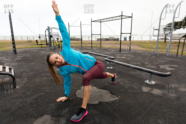 Front view of a sporty Caucasian woman with long dark hair exercising in an outdoor gym during daytime, leaning on the ground, stretching.