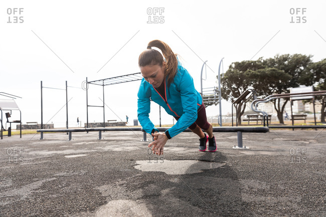 Front view of a sporty Caucasian woman with long dark hair exercising in an outdoor gym during daytime,  in mid air, doing push ups and clapping