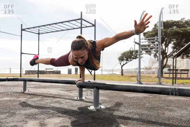 Front view of a sporty Caucasian woman with long dark hair exercising in an outdoor gym during daytime, balancing on one hand on a bench.