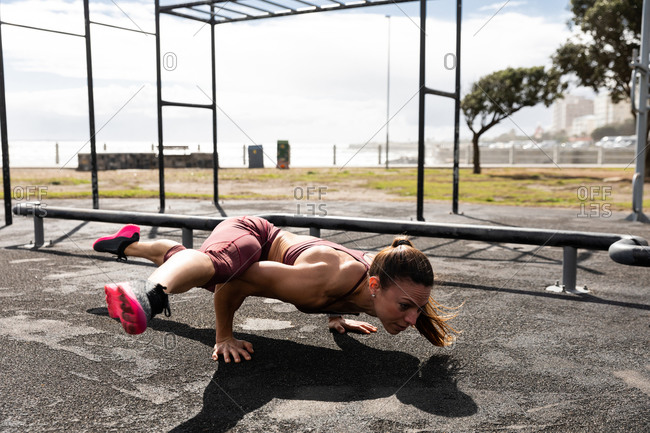 Side view of a sporty Caucasian woman with long dark hair exercising in an outdoor gym during daytime, balancing on her hands with her legs outstretched.