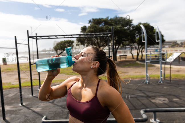 Front view of a sporty Caucasian woman with long dark hair exercising in an outdoor gym during daytime, drinking water from a bottle.