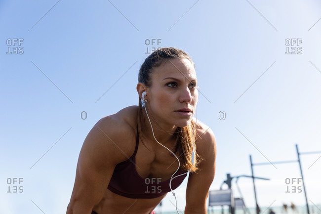 Low angle side view of a sporty Caucasian woman with long dark hair exercising in an outdoor gym during daytime, leaning on her knees with her headphones on.