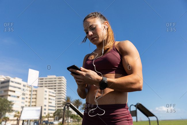 Low angle side view of a sporty Caucasian woman with long dark hair exercising in an outdoor gym during daytime, using her phone with her headphones on.