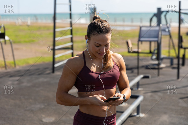 Front view of a sporty Caucasian woman with long dark hair exercising in an outdoor gym during daytime, using her phone with her headphones on.