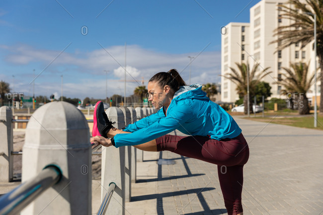 Side view of a sporty Caucasian woman with long dark hair exercising on a promenade by the seaside on a sunny day with blue sky, stretching her leg on a handrail.