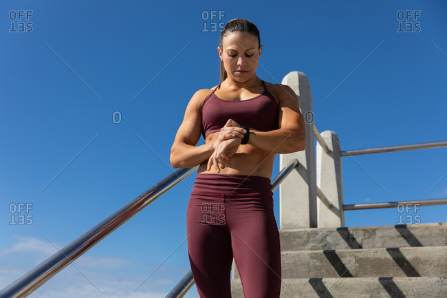 Low angle front view of a sporty Caucasian woman with long dark hair exercising on a promenade by the seaside on a sunny day with blue sky, checking her smartwatch.