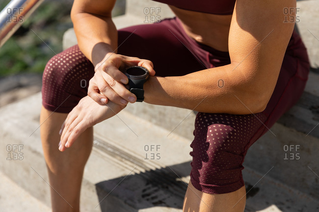 Side view mid section of a sporty Caucasian woman exercising on a promenade by the seaside on a sunny day, sitting on the stairs, looking at her smartwatch.