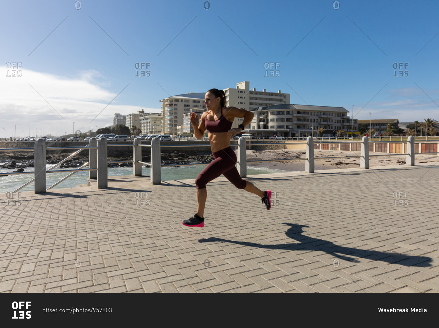 Side view of a sporty Caucasian woman with long dark hair exercising on a promenade by the seaside on a sunny day with blue sky, running.