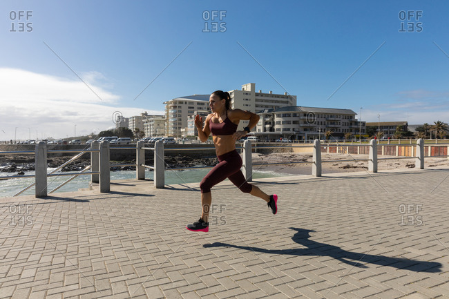 Side view of a sporty Caucasian woman with long dark hair exercising on a promenade by the seaside on a sunny day with blue sky, running.