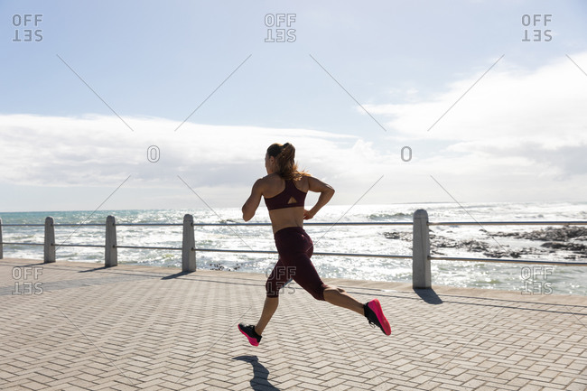 Side view of a sporty Caucasian woman with long dark hair exercising on a promenade by the seaside on a sunny day with blue sky, running.