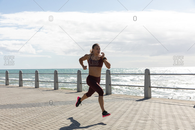 Side view of a sporty Caucasian woman with long dark hair exercising on a promenade by the seaside on a sunny day with blue sky, running.