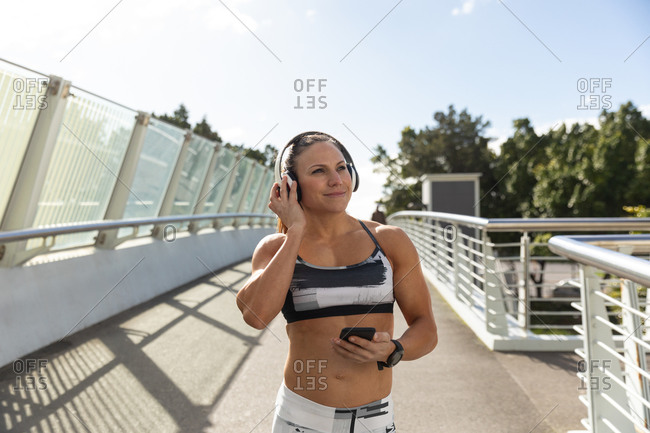 Front view of a sporty Caucasian woman with long dark hair exercising in an urban park on a sunny day with blue sky, enjoying the music with her headphones on.