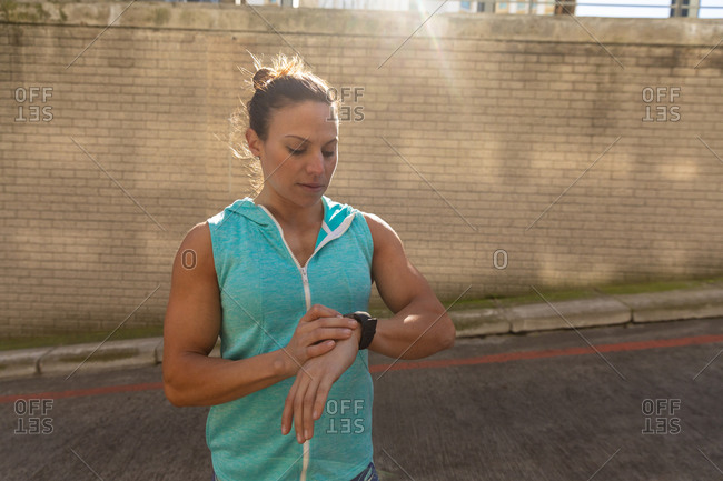 Front view of a sporty Caucasian woman with long dark hair exercising in the urban area on a sunny day, checking her smartwatch.