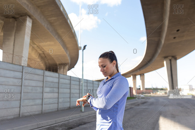 Side view of a sporty Caucasian woman with long dark hair exercising in the urban area on a sunny day, checking her smartwatch.