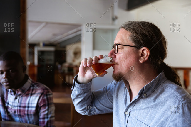 Caucasian man with long hair tasting beer in a microbrewery pub with an African American man sitting by a table in the background.