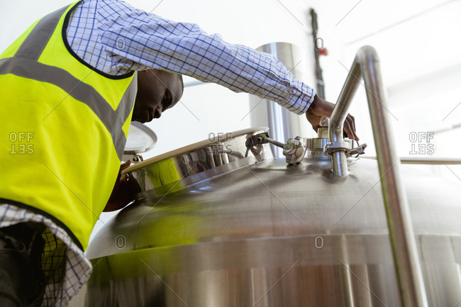Low angle view of an African American man working at a microbrewery, wearing a high visibility vest, inspecting beer and looking inside the tank.