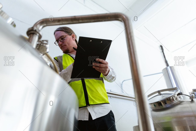 Low angle view of a Caucasian man wearing high visible vest, working in a microbrewery, holding a file and writing data while checking vats.