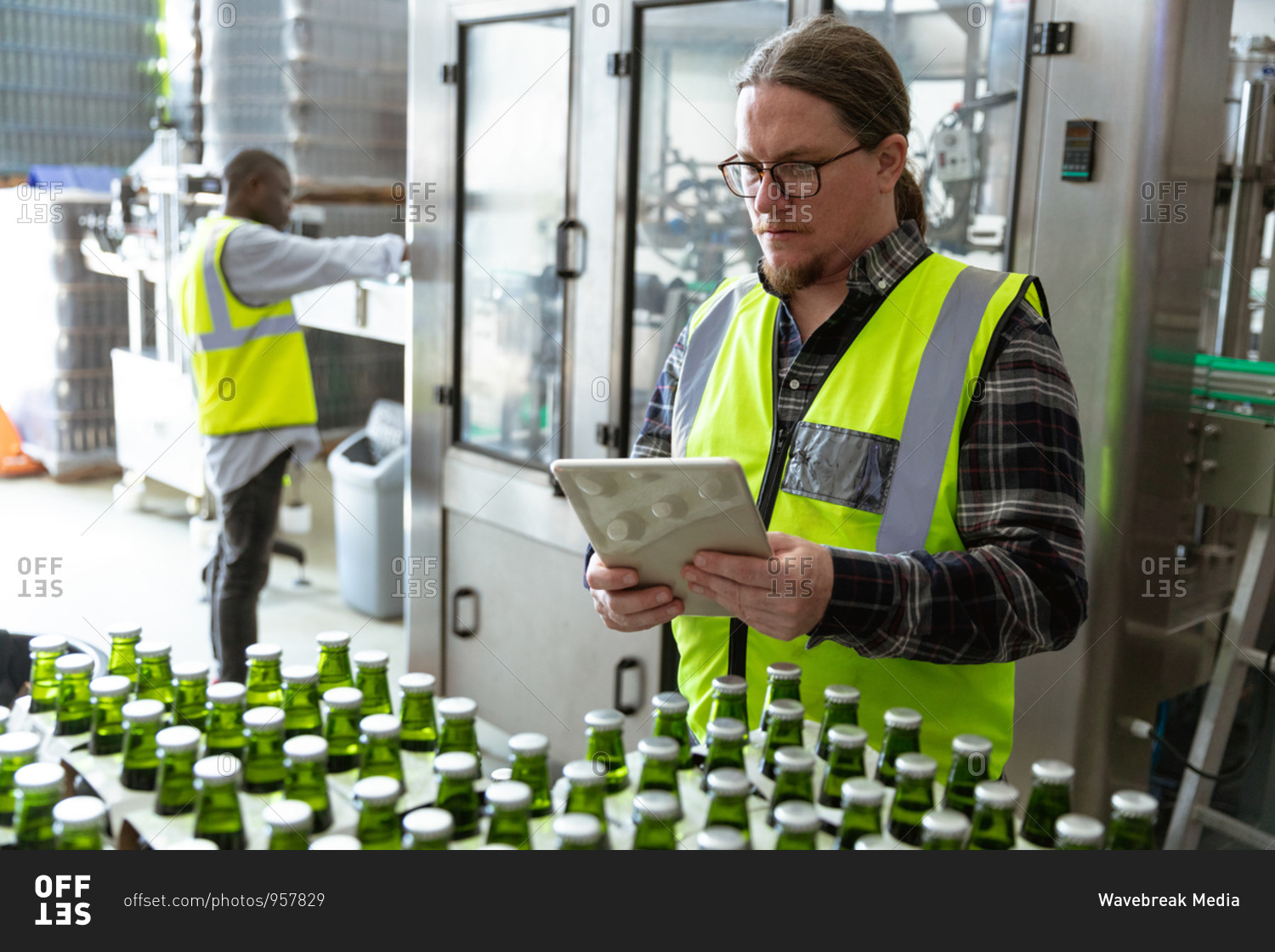 Caucasian man wearing high visibility vest, working in a microbrewery, using a tablet while checking bottles of beer prepared for delivery with an African American man working in the background.