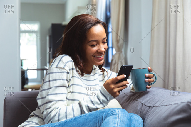 Mixed race woman enjoying her time at home, social distancing and self isolation in quarantine lockdown, sitting on a sofa, holding a mug and using a smartphone