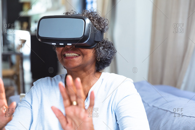 Senior mixed race woman enjoying her time at home, social distancing and self isolation in quarantine lockdown, sitting on a sofa, wearing vr goggles and touching virtual reality screen