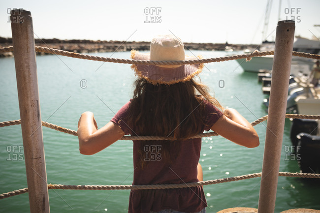 A teenage Caucasian girl, wearing a straw hat, enjoying her time on a promenade, on a sunny day,  sitting and leaning on a barrier, looking away