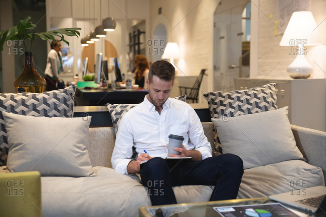 Caucasian male business creative working in a casual modern office, sitting on a sofa drinking a coffee and making notes, with colleagues working in the background