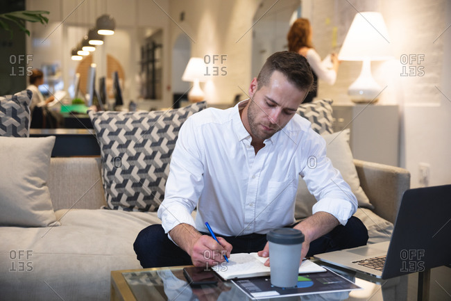 Caucasian male business creative working in a casual modern office, sitting on a sofa and making notes, with colleagues working in the background