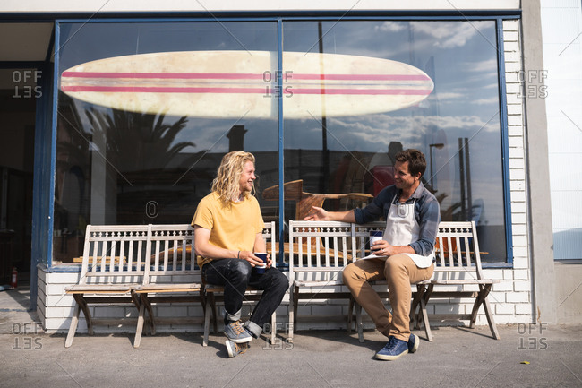 Two Caucasian male surfboard makers taking a break during work in their studio, sitting on a bench and interacting while drinking takeaway coffee and having a snack.