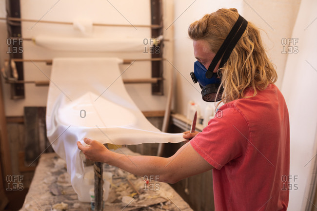 Caucasian male surfboard maker wearing a breathing face mask, working in his studio, inspecting a surfboard covered with a white piece of cloth.