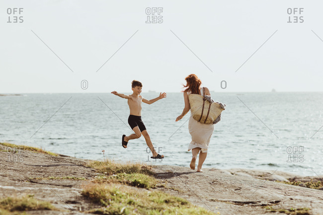 Rear view of woman with bag while son jumping against sea during sunny day