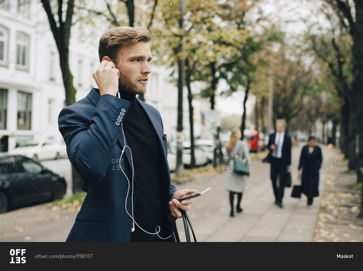 Businessman looking away while holding in-ear headphones in city