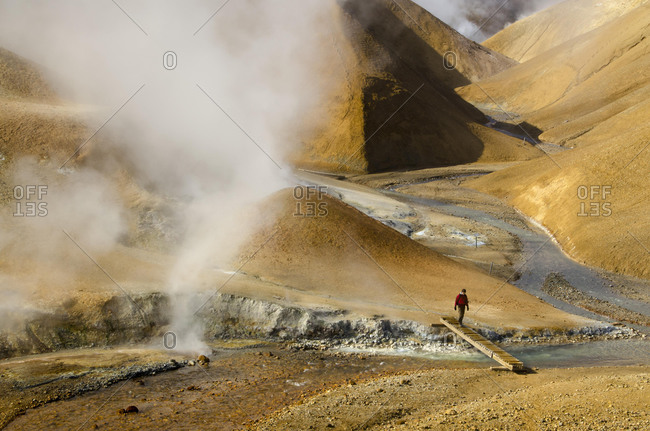 Hiker on a bridge over a steaming creek on a hiking trail, hot springs and the snow-capped Rhyolite Mountains, Hveradallir high temperature area, Kerlingarfjoell, highlands, Iceland, Europe