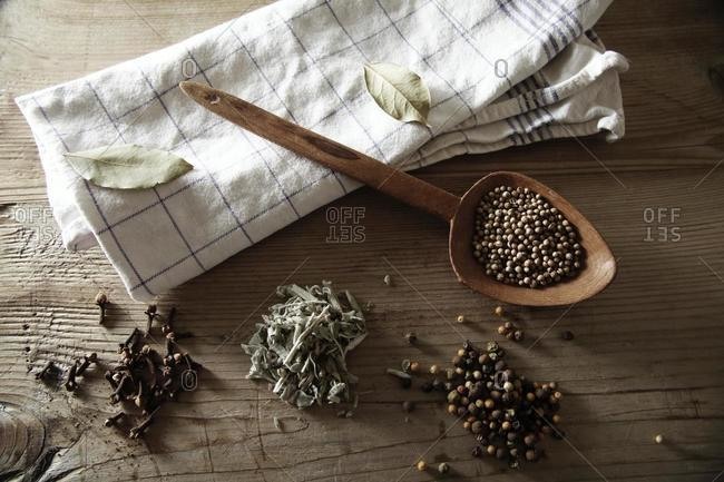 Coriander (Coriandrum sativum), Peppercorns (Piper nigrum), Sage (Salvia) and dried Cloves (Syzygium aromaticum) with a wooden spoon and a kitchen towel on a rustic wooden surface