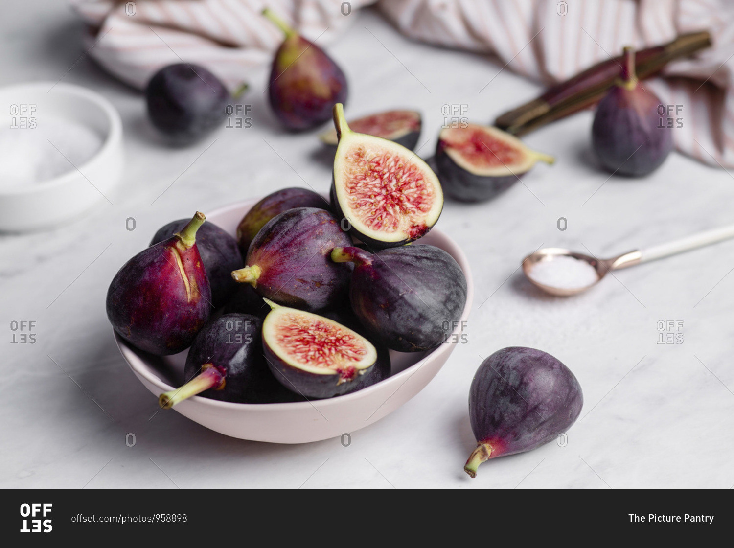 Close up of a bowl of ripe, purple figs on a white marble countertop surrounded by a striped napkin, and a rose gold spoonful of sugar