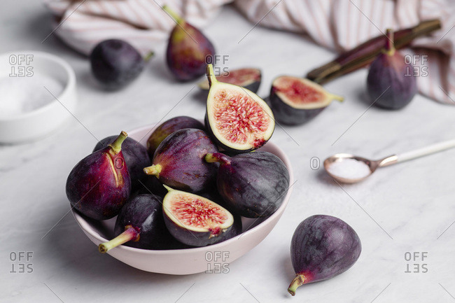 Close up of a bowl of ripe, purple figs on a white marble countertop surrounded by a striped napkin, and a rose gold spoonful of sugar