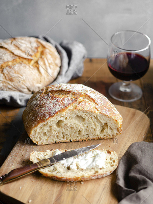 A homemade loaf of bread, sliced and buttered on a wood cutting board sitting atop a rustic table with a glass of red wine in the background