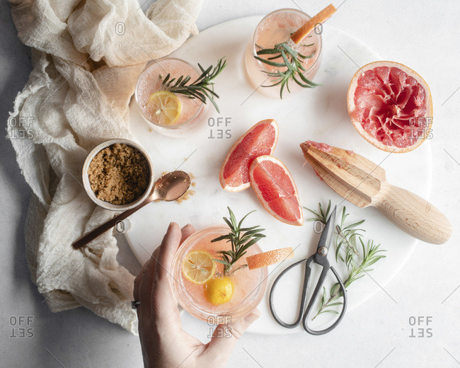 Overhead shot a hand reaching for a glass of grapefruit cocktail on a round platter with rosemary and grapefruit slices and a cup of brown sugar with a rose gold spoon