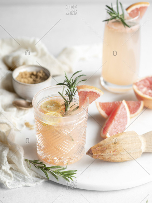 Old fashioned drinking glass filled with a grapefruit juice cocktail and garnished with lemon and rosemary on a white tray with a wooden hand juicer