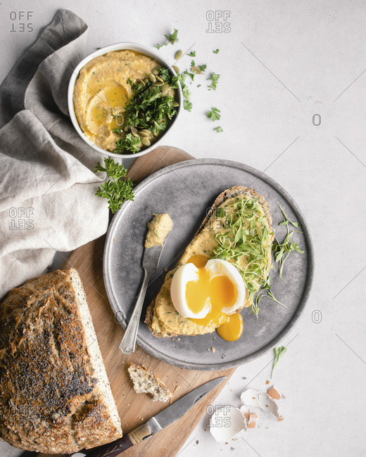 A slice of bread topped with hummus, herbs, and a soft boiled egg, surrounded by the cut loaf, eggshells, a napkin, and a small bowl of hummus on a white countertop
