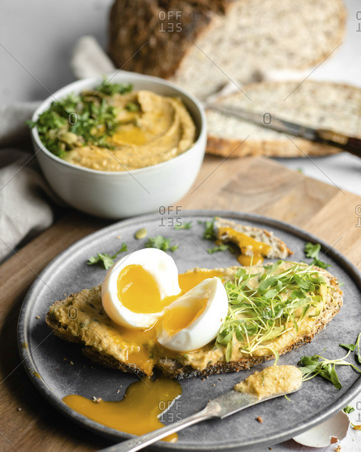 A soft boiled egg, cut in half on a thin slice of bread covered with hummus and greens on a silver plate sitting on a wood cutting board, with a bowl of hummus and the loaf of sliced bread in the blurred background